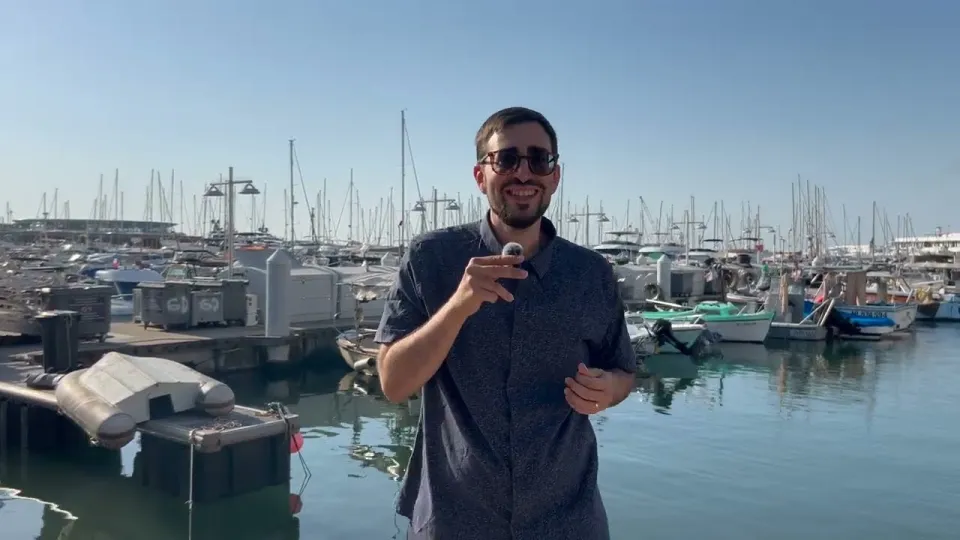 Mickael speaking to camera on the quayside at Cannes, boats and masts behind him